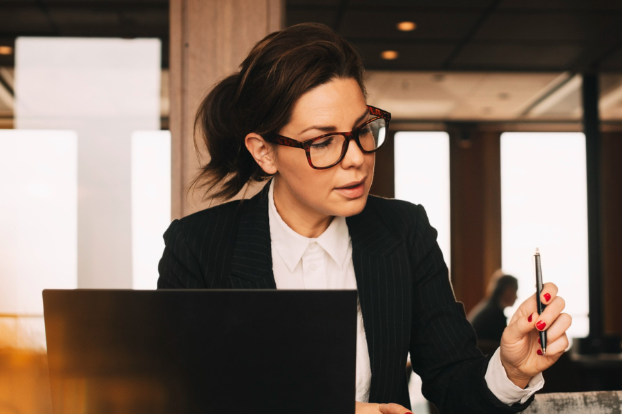A professional woman wearing glasses and a pinstripe blazer is seated at a desk with a laptop